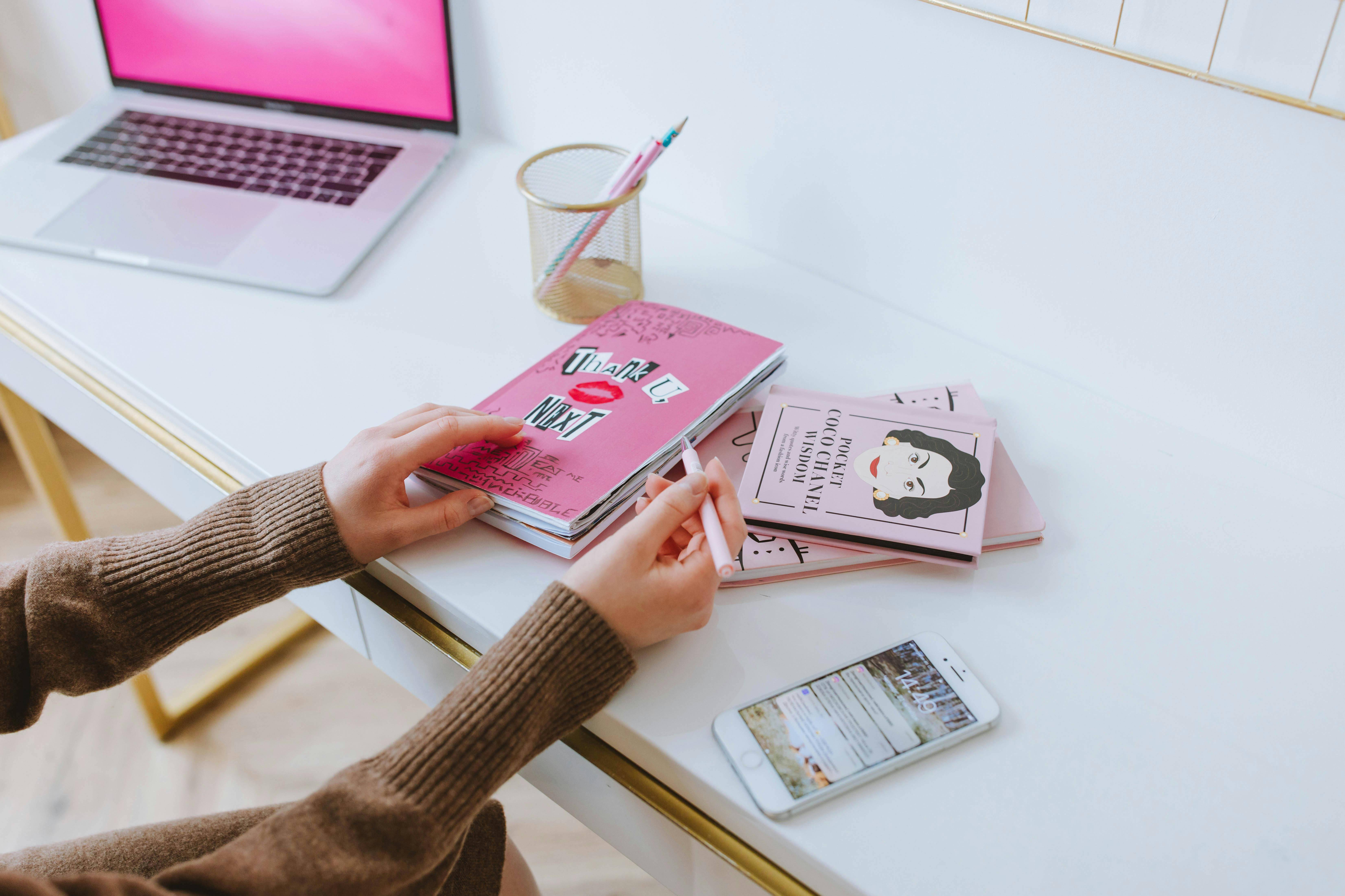 A person wearing a brown sweater is sitting at a white desk, holding a pink pen and interacting with a pink notebook that has a sticker reading 
