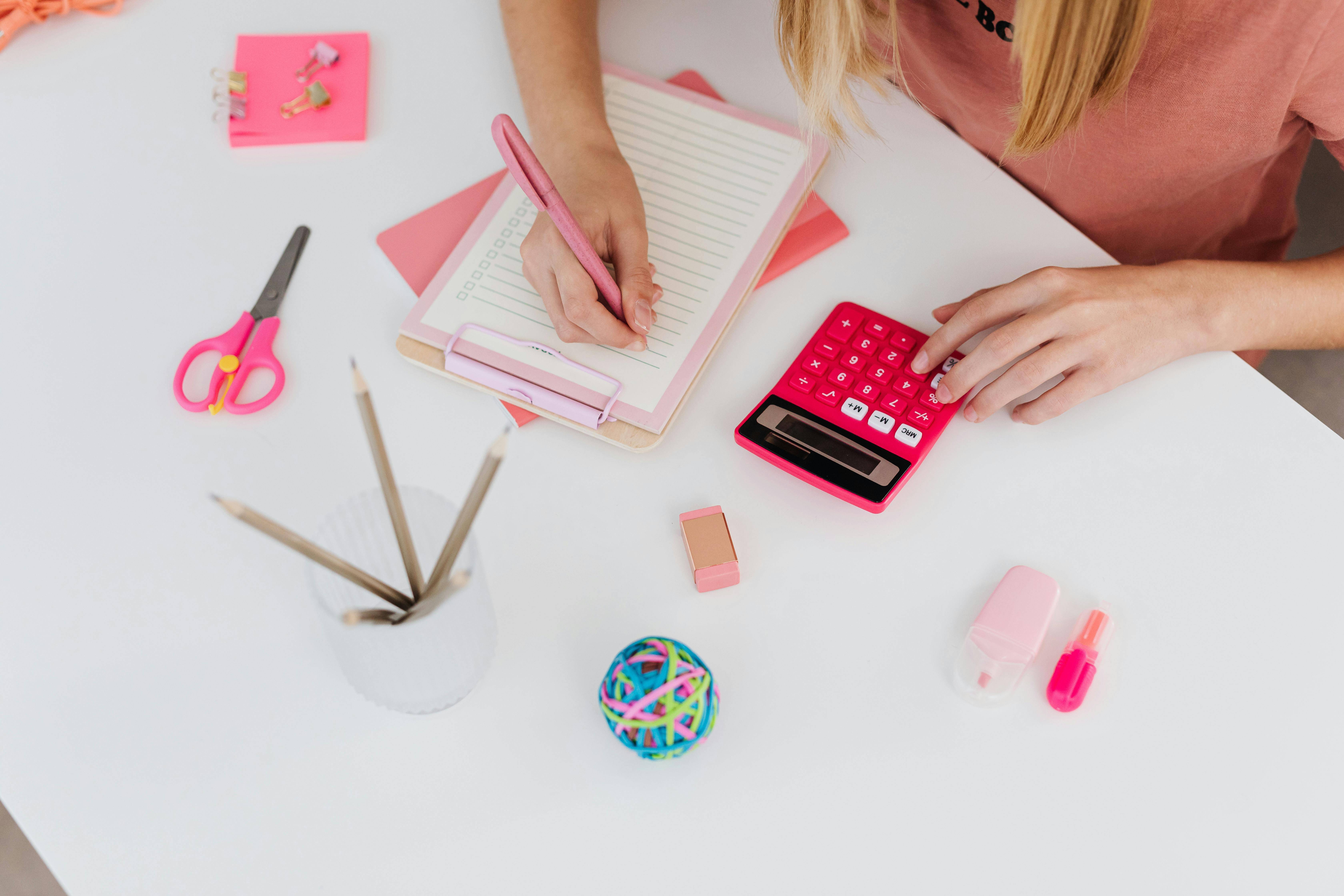 A person wearing a pink t-shirt is working at a white desk, using a pink calculator with one hand while writing on a clipboard with the other. The desk is neatly organized with various office supplies, including pink-handled scissors, a cup filled with pencils, a rubber band ball, an eraser, and a small pink correction tape dispenser. The scene has a cohesive pink and white color scheme, creating a bright and aesthetically pleasing workspace.